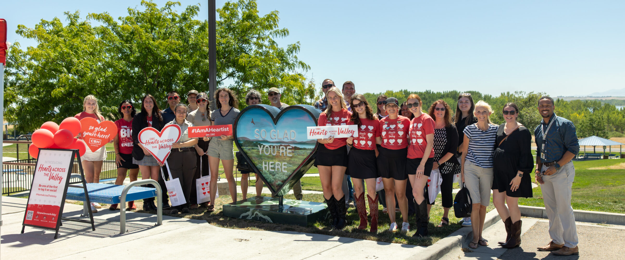 Group photo at the Hearts Across the Valley art event in Caldwell, Idaho, featuring community members and CBH Homes team around the “So Glad You’re Here” heart installation at Mallard Park.