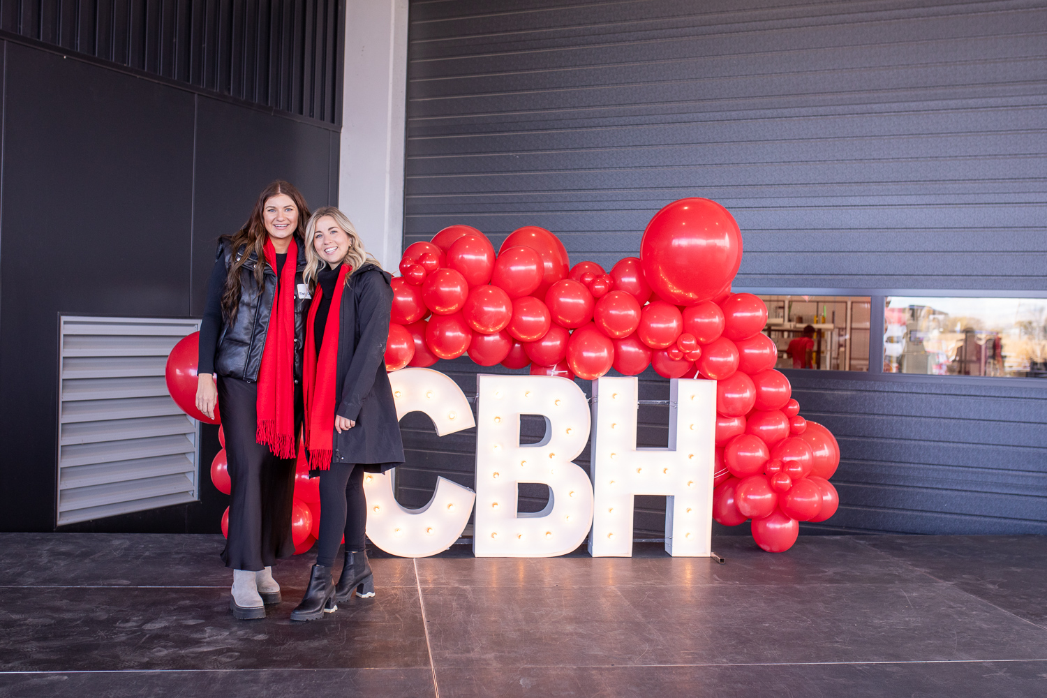 Two smiling women stand in front of large illuminated ‘CBH’ letters decorated with bright red balloons at an event. Both are wearing black coats and red scarves, posing together on a stage area outside a building.
