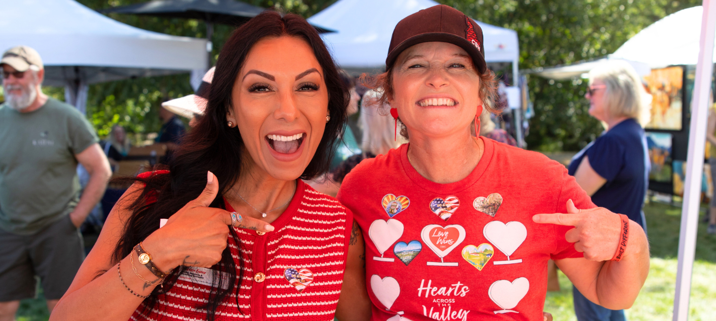 Two women smile and point to their Hearts Across the Valley apparel while standing at a Heart Unveiling. One wears a red and white patterned vest with a heart pin, and the other wears a red Hearts Across the Valley T-shirt covered in heart graphics. Vendor tents and attendees can be seen in the background, highlighting the community spirit of the event.