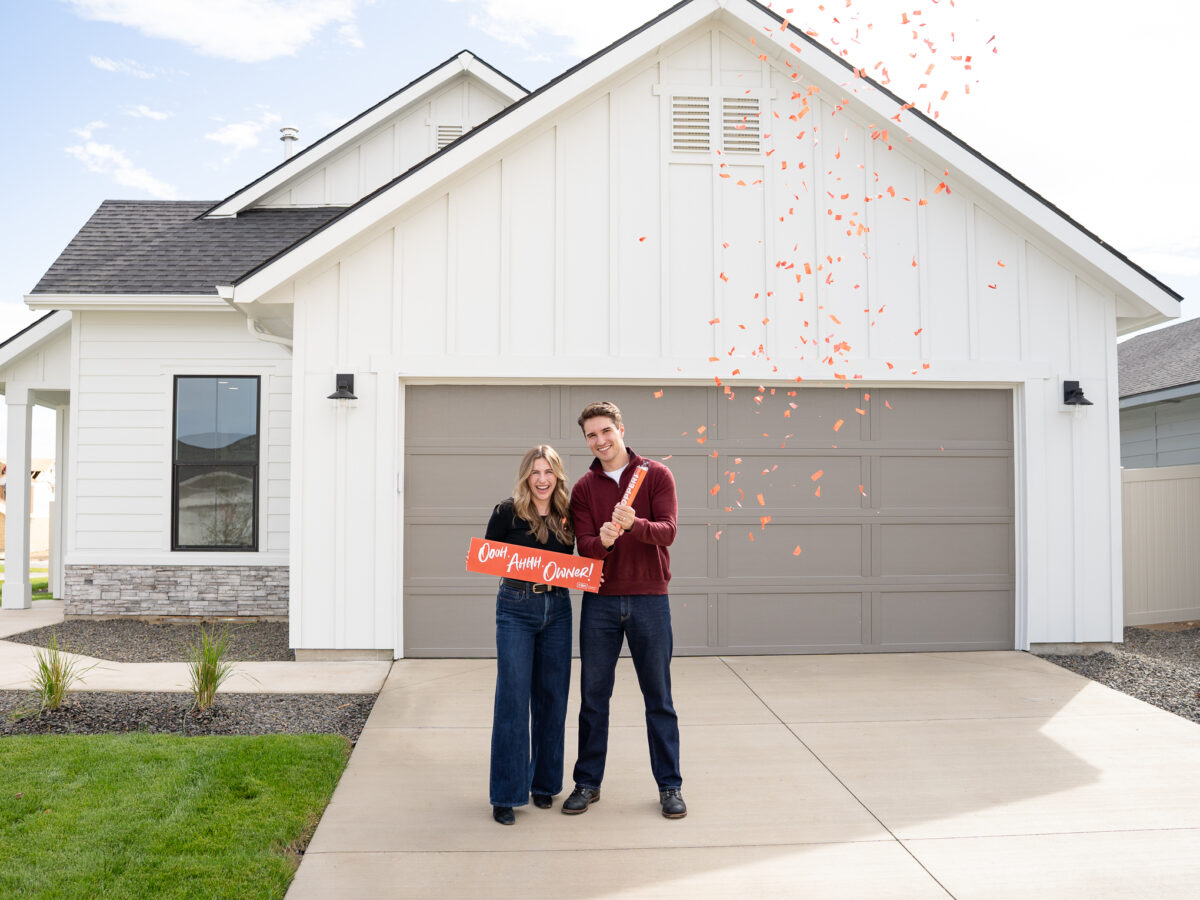 Happy new homeowners celebrating with red confetti in front of their new CBH Homes house in Idaho, holding an “Ooh Ahhh Owner!” sign.