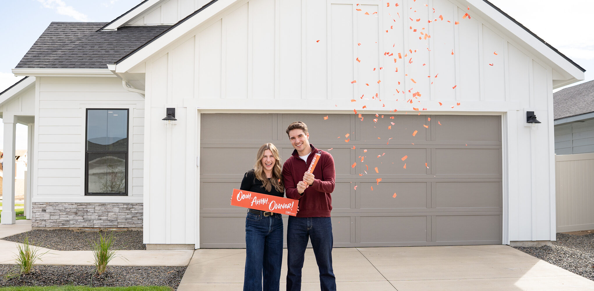 Happy new homeowners celebrating with red confetti in front of their new CBH Homes house in Idaho, holding an “Ooh Ahhh Owner!” sign.