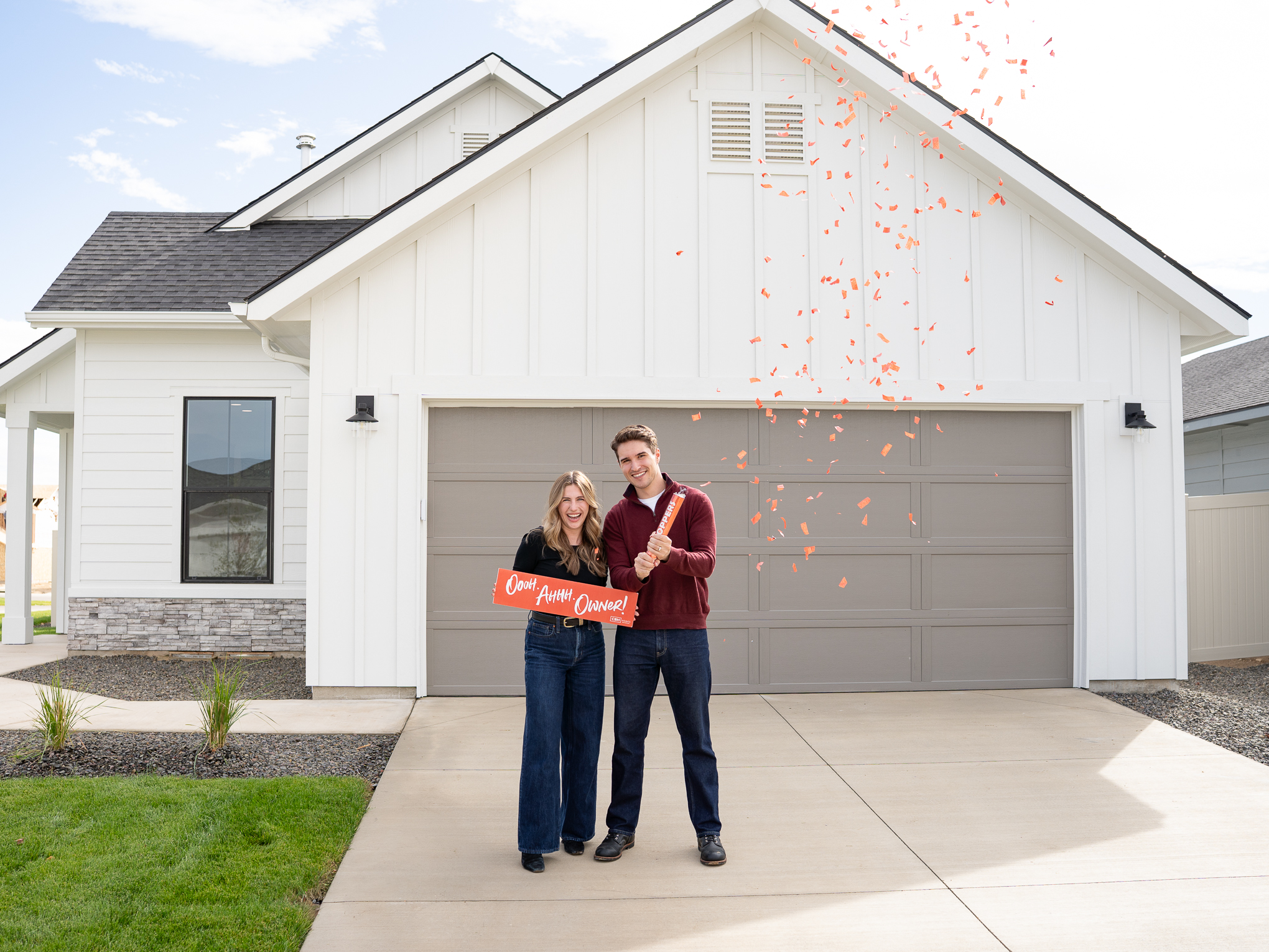 Happy new homeowners celebrating with red confetti in front of their new CBH Homes house in Idaho, holding an “Ooh Ahhh Owner!” sign.