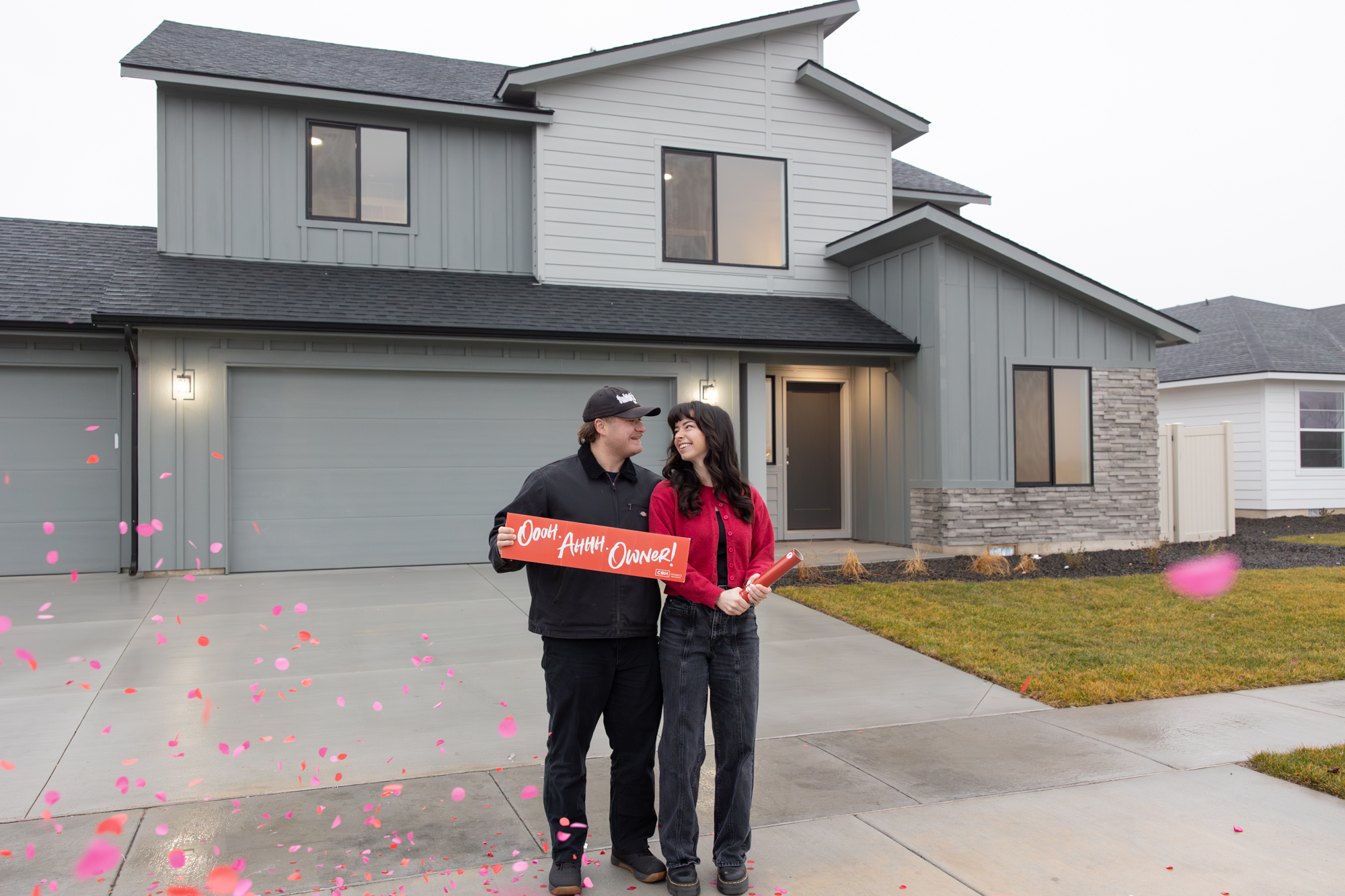 Couple holding an “Ooo! Ahh! Owner!” sign with pink confetti in front of a newly built two-story CBH home with a three-car garage in an Idaho neighborhood.