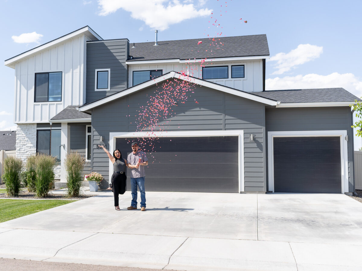 Couple celebrating in front of a newly built gray two-story CBH home with a three-car garage as pink confetti falls, marking their new home purchase in an Idaho neighborhood.