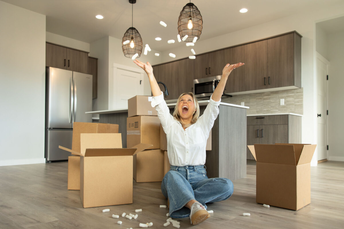 Excited new homeowner sitting on the floor of a modern kitchen surrounded by moving boxes, tossing packing peanuts in the air in celebration of moving into a new home.