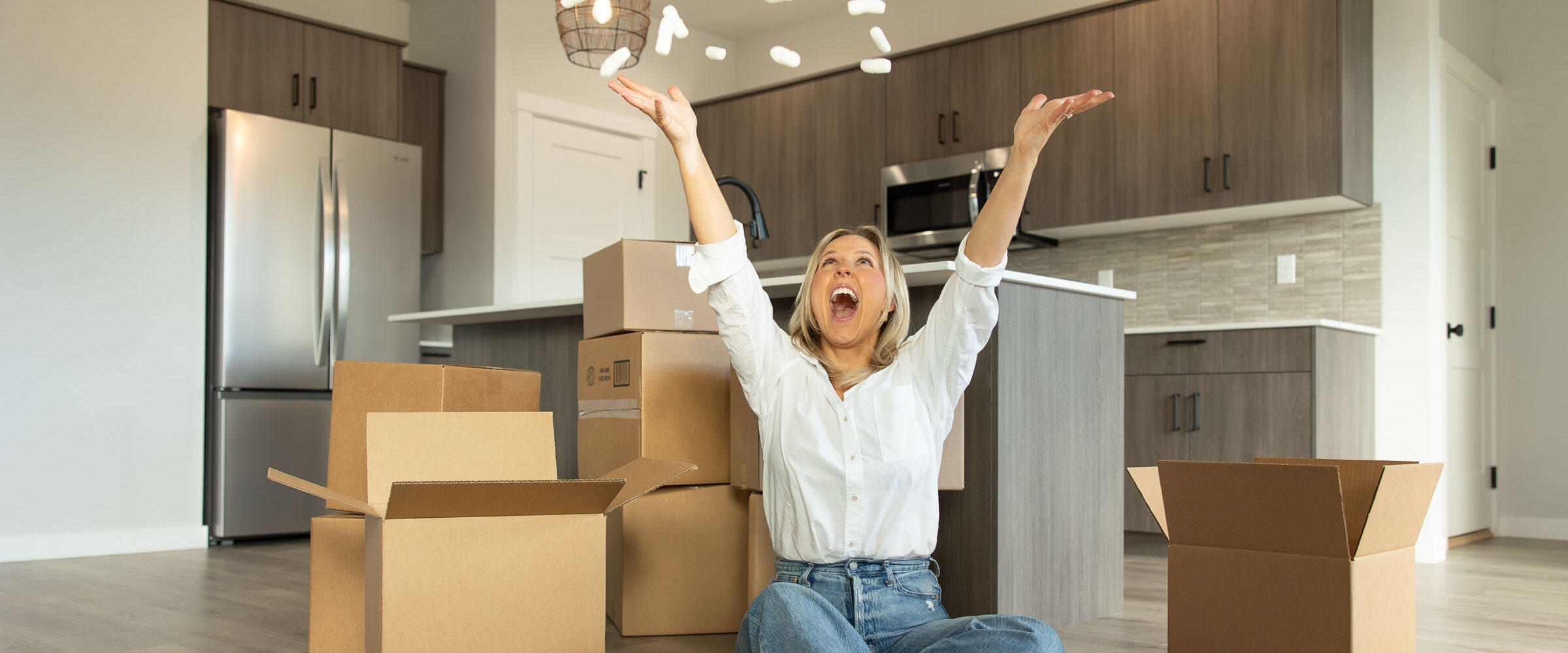 Excited new homeowner sitting on the floor of a modern kitchen surrounded by moving boxes, tossing packing peanuts in the air in celebration of moving into a new home.