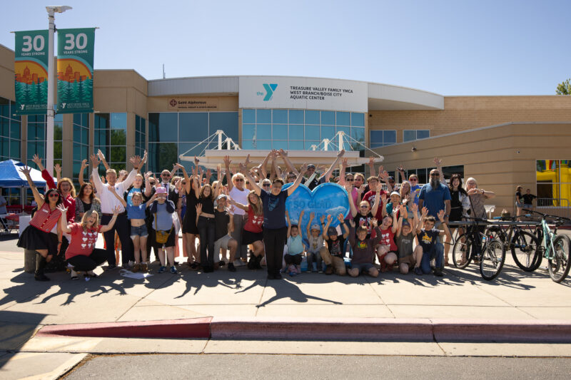 A large, cheerful group poses with arms raised outside the West Boise YMCA, holding the blue "SO GLAD YOU'RE HERE" heart sculpture. "30 Years Strong" banners hang nearby on a sunny day.