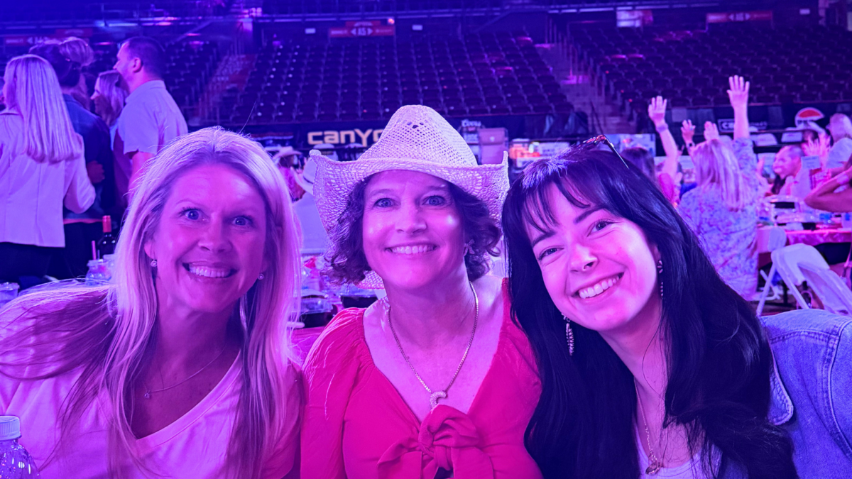 Three women smiling at the Pink on the Dirt luncheon inside Ford Idaho Center during the 2026 Snake River Stampede, with the arena lit in pink and purple