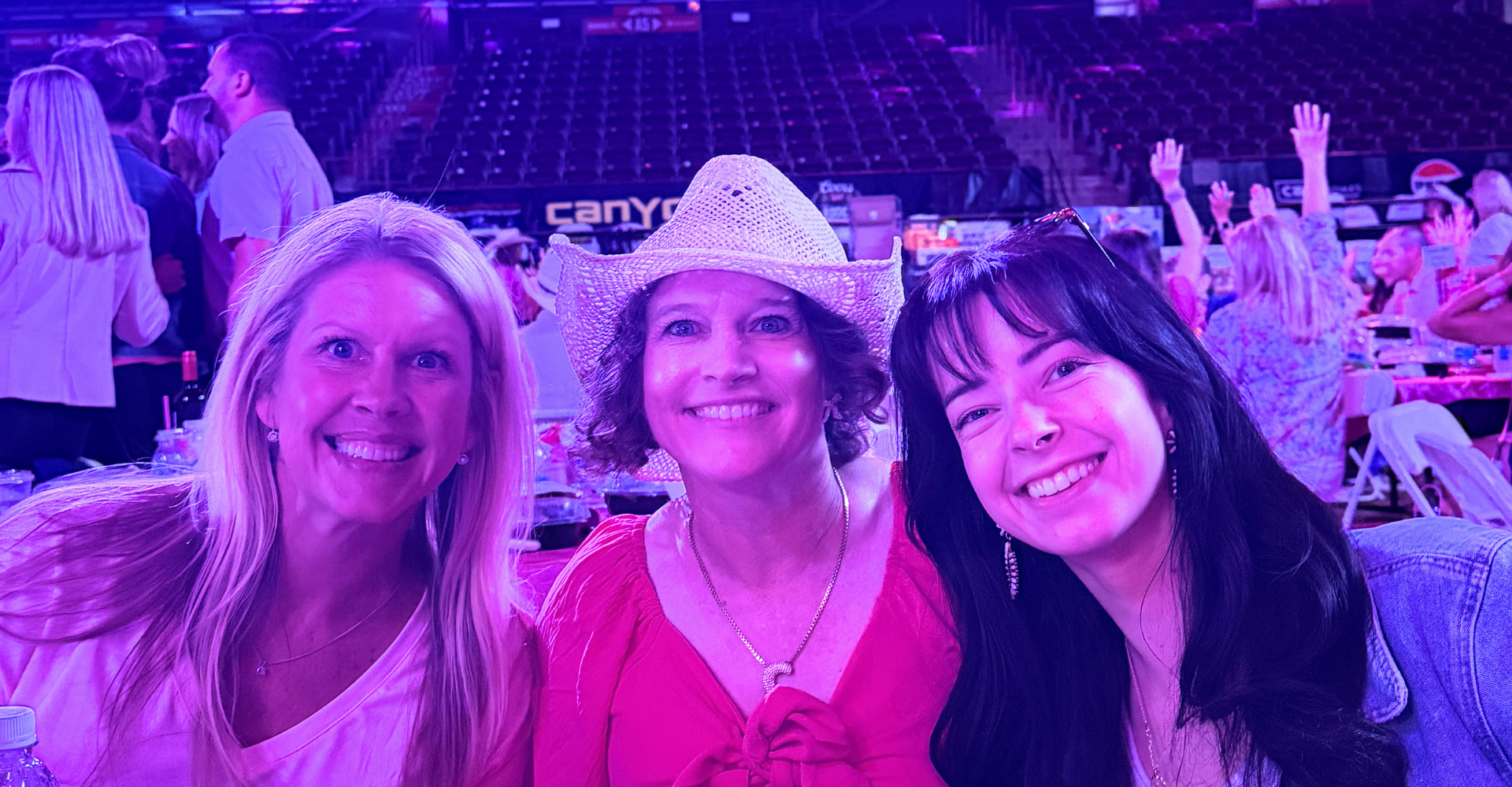 Three women smiling at the Pink on the Dirt luncheon inside Ford Idaho Center during the 2026 Snake River Stampede, with the arena lit in pink and purple