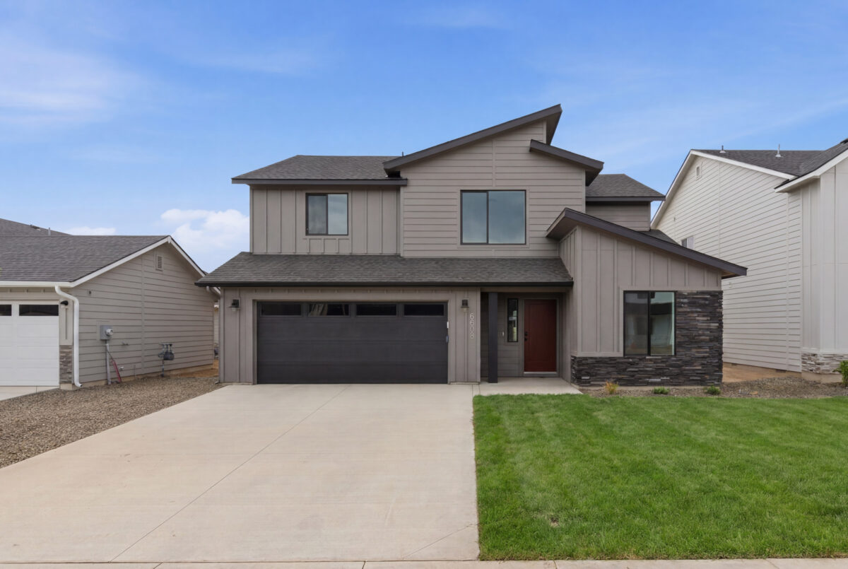 Modern two-story CBH home with warm gray board-and-batten siding, dark stacked stone accents, charcoal garage door, and a bold red front door in the Treasure Valley