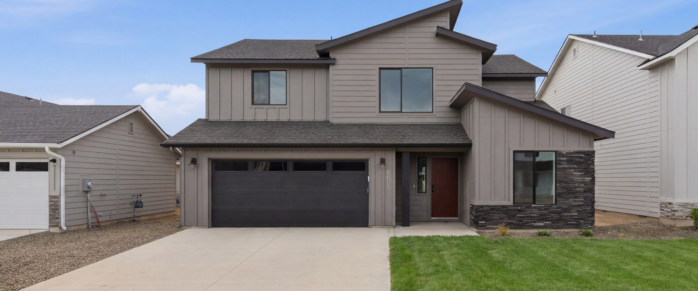 Modern two-story CBH home with warm gray board-and-batten siding, dark stacked stone accents, charcoal garage door, and a bold red front door in the Treasure Valley